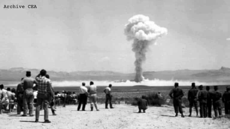 Photo des essais nucléaires de la France en Algérie. Plusieurs personnes debout face à un champignon nucléaire au fond. Gerboise bleue. Région de Reggane