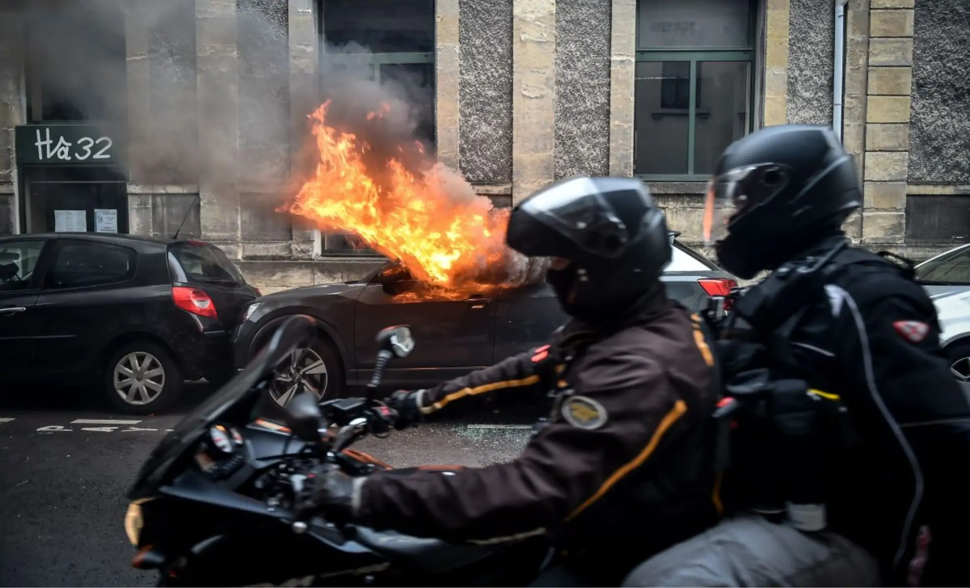 personnes à moto passant devant une voiture en feu, en France