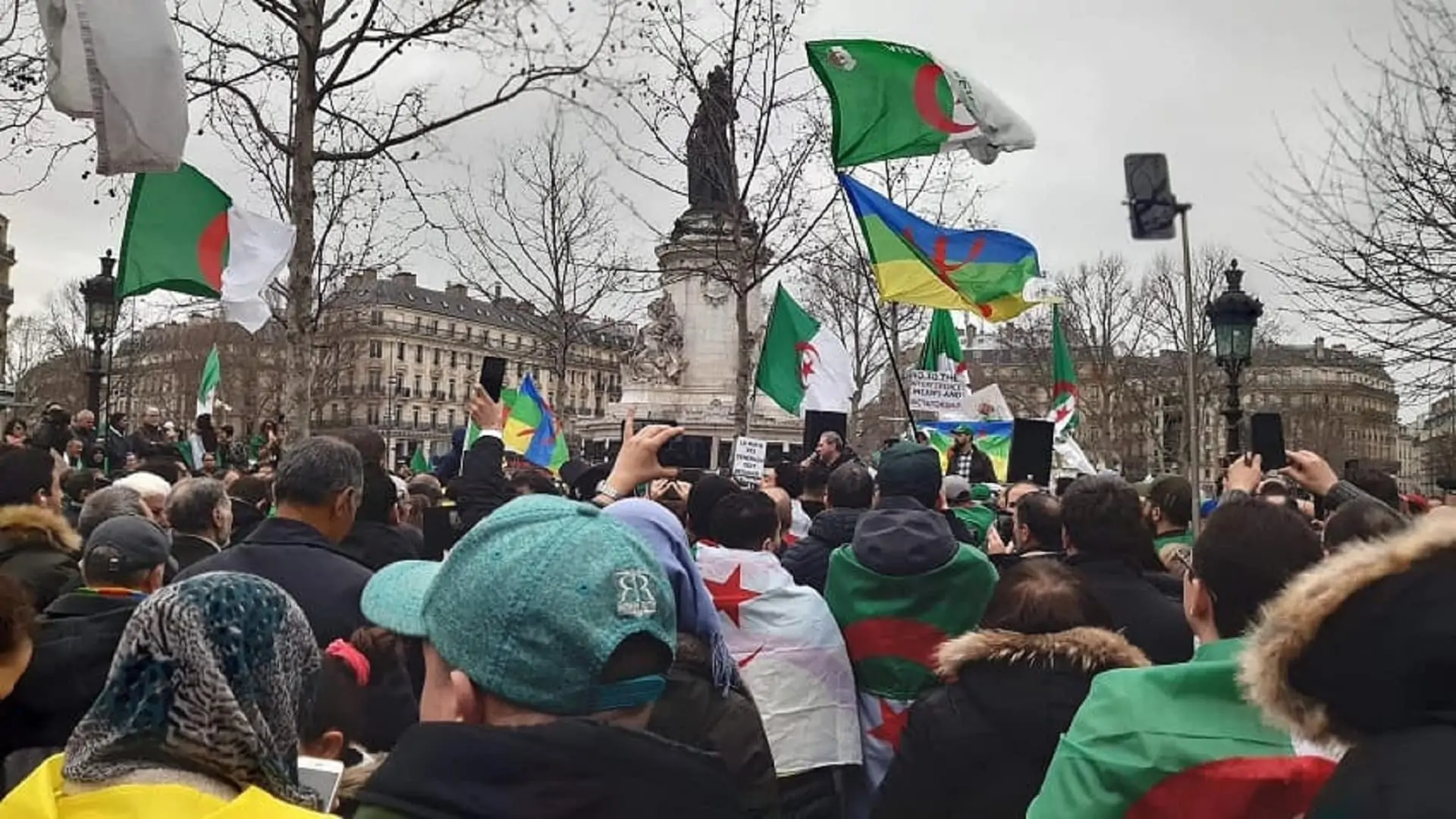 Photo d'Algériens de France manifestant à la Place de la République à Paris. Drapeaux algériens et berbères brandis. Statue au centre de l'image.
